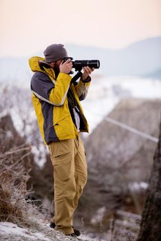 Young cheerful male photographer taking photographs of winter forest with digital camera at mountains  Young man shooting photos outdoors