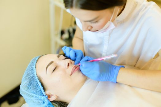 Skilled female cosmetologist shaping lips with pencil for young girl before microblading. Concept of permanent makeup artist and beauty salon.