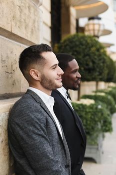 Afro american and european gays standing near building and wearing suits. Concept of lgbt and walking in city.