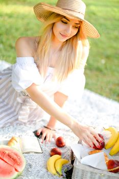 Young woman in hat eating fruits and lying on plaid with book, grass in backrground. Concept of healthy food, picnic and summer season.