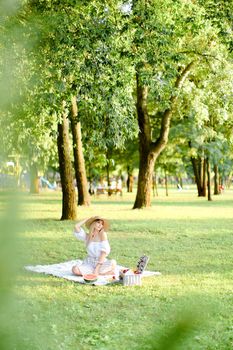 Young caucasian blonde woman in hat sitting on plaid with fruits in garden. Concept of summer picnic in park, nature and free time.