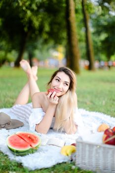 Young cute woman lying on plaid in park, reading book near watermelon and keeping apple. Concept of leisure time, summer vacations and picnic, healthy food.