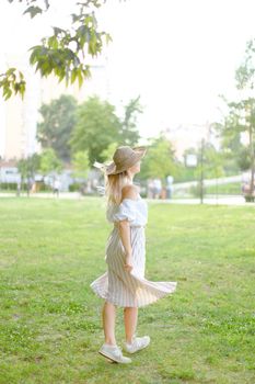 Back view of young girl wearing hat and dress, standing in park on grass. Concept of summer season fashion and female person.