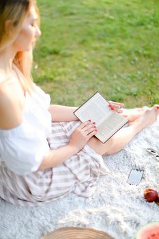 Young girl reading novel and sitting on plaid near fruits in park, focus on book. Concept of having summer picnic and leisure time.