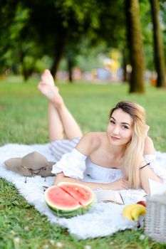 Young blonde woman lying on plaid in park, reading book near watermelon and hat. Concept of leisure time, summer vacations and picnic.