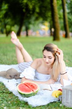 Young european girl lying on plaid in park near watermelon and reaing book. Concept of picnic and resting in open air on weekends.