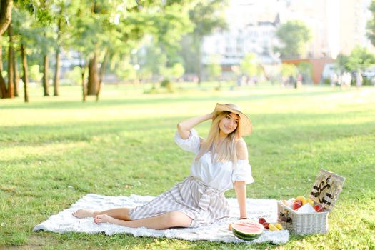 Young blonde caucasian woman in hat sitting in park on plaid near fruits, grass in background. Concept of summer picnic, resting on nature and healthy food.