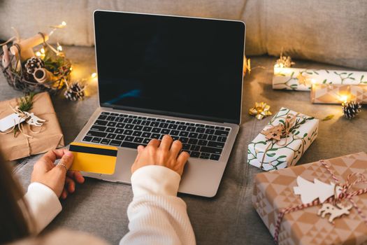 Close up of woman hands with credit card, gifts, and mock up screen laptop. Online shopping at Christmas holidays. Cropped female sit on couch with natural eco presents and decor. Merry Christmas.