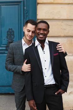 Two boys, caucasian and afro american, wearing suits standing near building and hugging. Concept of gays and lgbt.