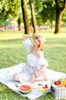 Young european girl in hat sitting in park on plaid near fruits, grass and trees in background. Concept of summer picnic, resting on nature and healthy food.