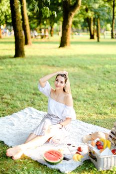 Young european caucasian girl having picnic on plaid and sitting in park with fruits. Concept of resting in open air, leisure time and summer season