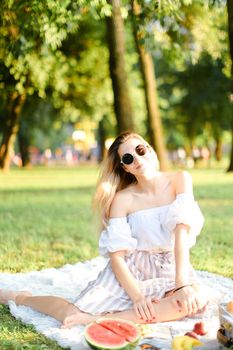 Young caucasian nice woman in sunglasses having picnic on plaid and sitting in park with fruits. Concept of resting in open air, leisure time and summer season