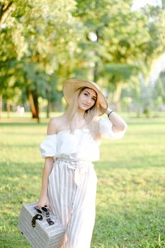 Young beautiful girl walking in garden and keeping bag, sunglasses and hat. Concept of walking in park and summer fashion.