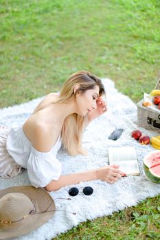 Young caucasian blonde woman reading book and lying on plaid near fruits and hat, grass in background. Concept of summer picnic and resting on weekends in open air.