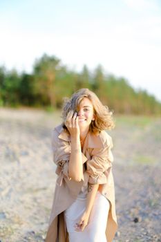 Smiling blonde woman wearing coat standing on sand with trees in background. Concept of happiness, youth and fashion.