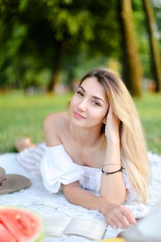 Young caucasian woman lying on plaid near watermelon in park and reading book. Concept of picnic on nature, summer vacations and leisure time.