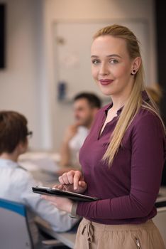 Blonde businesswoman in casual  clothes working on tablet computer at modern startup business office interior. Young people group on team meeting blured  in background