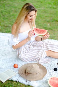 Young caucasian girl eating watermelon, sitting on plaid near fruits and hat with grass in background. Concept of summer picnic and resting on weekends in open air.