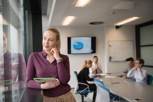 Blonde businesswoman in casual  clothes working on tablet computer at modern startup business office interior. Young people group on team meeting blured  in background