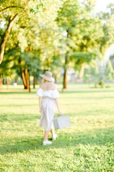 Blurry image of young girl wearing hat walking in garden with bag in sunny day. Concept of summer season, fashion and female person walking in park.