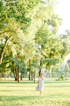 Young caucasian girl wearing hat walking in garden with bag in sunny day. Concept of summer season, fashion and female person walking in park.