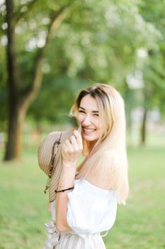 Young smiling girl wearing dress, standing in park and keeping hat. Concept of summer season and fashion.