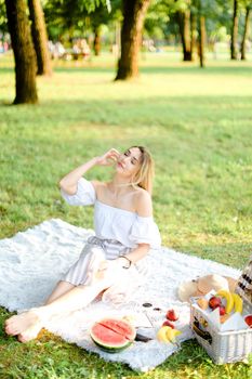 Young caucasian blonde girl having picnic on plaid and sitting in park with fruits. Concept of resting in open air, leisure time and summer season