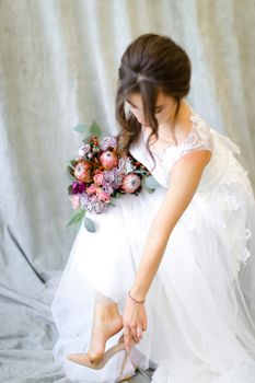 Young bride with bouquet of flowers putting on shoes at studio. Concept of bridal photo session and wedding.