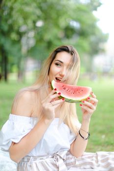 Young european girl eating watermelon and wearing dress, grass in background. Concept of summer photo session, picnic.