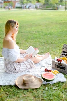 Young blonde girl reading book and sitting on plaid near fruits in park. Concept of having summer picnic and leisure time.