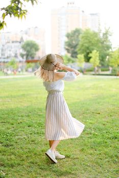 Back view of young woman wearing hat and dress, standing in park on grass. Concept of summer season fashion and female person.