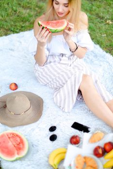 Caucasian blonde woman sitting on plaid near fruits and hat, eating watermelon, grass in background. Concept of summer picnic and resting on weekends in open air.