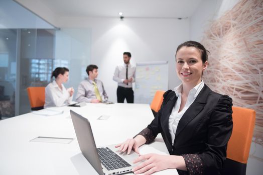 young business woman on meeting usineg laptop computer, blured group of people in background at  modern bright startup office interior taking notes on white flip board and brainstorming about plans and ideas