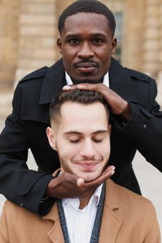 Afro american boy holding caucasian guy head by hands, wearing white shirt. Concept of stylish boy and haircut. Same sex couple, lgbt gays