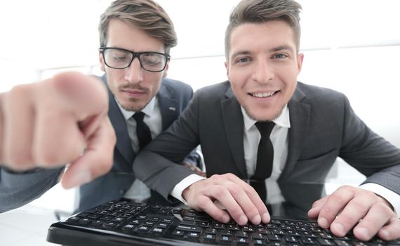 two young businessman are looking into the camera while sitting in the office. One shows the index finger