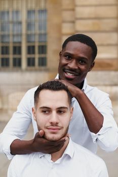 Afro american boy holding caucasian guy head by hands, wearing white shirt. Concept of stylish boy and haircut. Same sex couple, lgbt gays