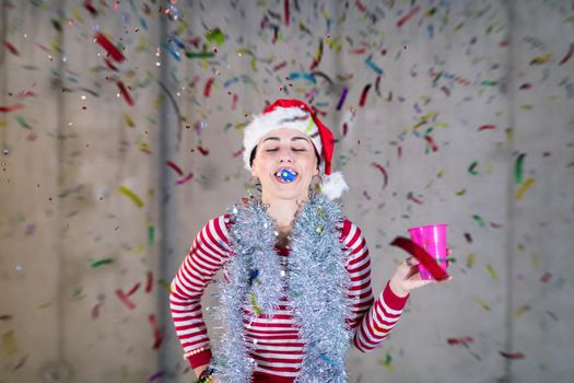 young happy casual business woman wearing a red hat and blowing party whistle while having new years confetti party in front of concrete wall