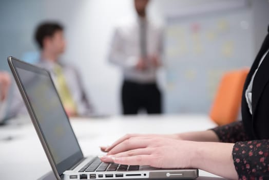 young business woman on meeting usineg laptop computer, blured group of people in background at  modern bright startup office interior taking notes on white flip board and brainstorming about plans and ideas