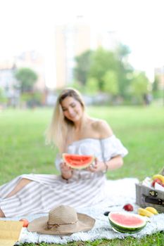 Young woman sitting on plaid near fruits and hat, eating watermelon, grass in background. Concept of summer picnic and resting on weekends in open air.