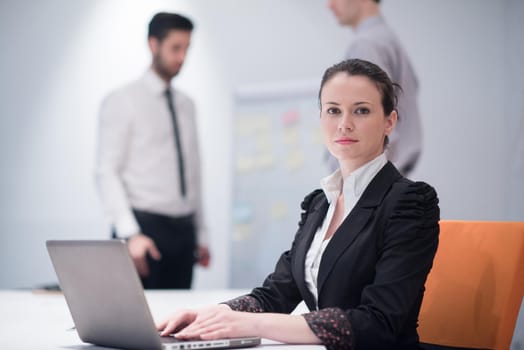 young business woman on meeting usineg laptop computer, blured group of people in background at  modern bright startup office interior taking notes on white flip board and brainstorming about plans and ideas