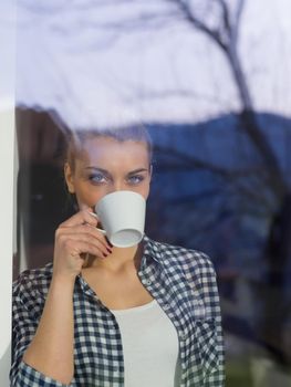 beautiful young woman drinking morning coffee by the window in her home