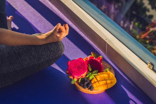 hand of a woman meditating in a yoga pose, sitting in lotus with fruits in front of her dragon fruit, mango and mulberry.