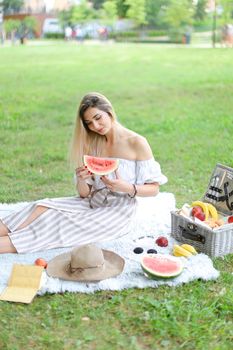 Young girl sitting on plaid near fruits and hat, eating watermelon, grass in background. Concept of summer picnic and resting on weekends in open air.