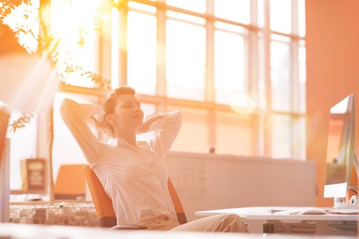 Happy young business woman relaxing and geting insiration while working on desktop computer at modern bright  starup office interior. Morning sunrise or sunset with sun flare in background.