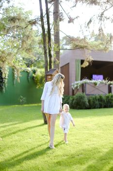 Young mom walking with child on grass in yard. Concept of motherhood and kids, summer season.