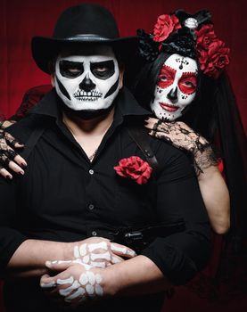 beautiful woman with a sugar skull makeup with a wreath of flowers on her head and a skeleton man in a black hat. Couple on dark red background