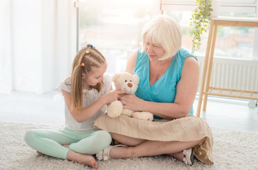 Cute little girl playing plush teddy bear toy with mature woman sitting on floor in modern apartment