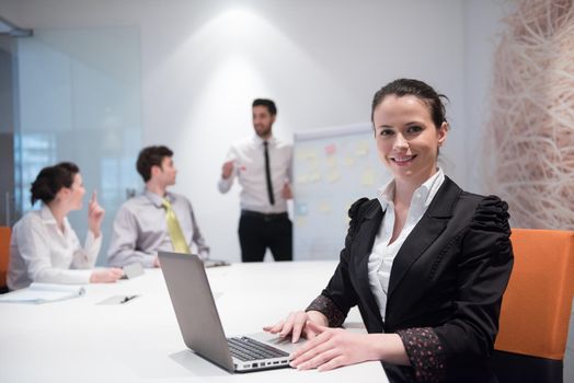 young business woman on meeting usineg laptop computer, blured group of people in background at  modern bright startup office interior taking notes on white flip board and brainstorming about plans and ideas