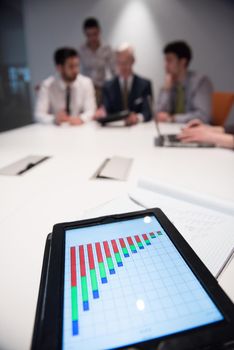 Close up of tablet touchpad computer with focus on business analytics and progress chart document. Business people group on meeting in background