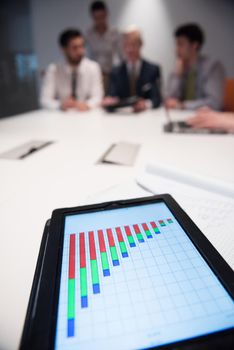 Close up of tablet touchpad computer with focus on business analytics and progress chart document. Business people group on meeting in background